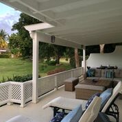 A covered porch area features a combination of outdoor seating and a coffee table, arranged for relaxation. Lush green surroundings are visible beyond the railing, offering a peaceful view of the landscape. The space is well-lit by natural light filtering through the overhead structure.
