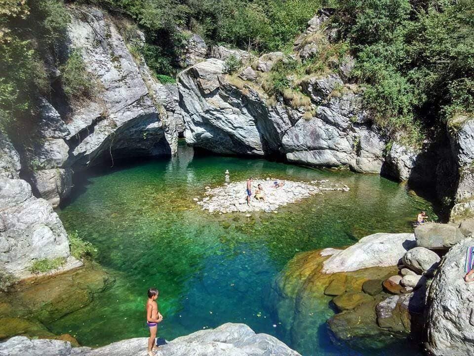 A natural pool is surrounded by large rocks and lush greenery. Clear turquoise water reflects sunlight, creating a serene environment. A small sandy area in the center serves as a gathering point, while individuals can be seen enjoying the water and the beauty of the surrounding landscape.