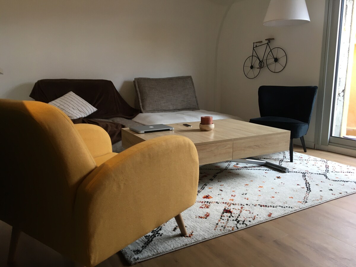 A furnished living area is highlighted by a yellow armchair and a black chair, both positioned around a wooden coffee table. A light-colored sofa bed is visible in the background, complementing the cozy space. A decorative bicycle wall piece adds an artistic touch to the simple decor.