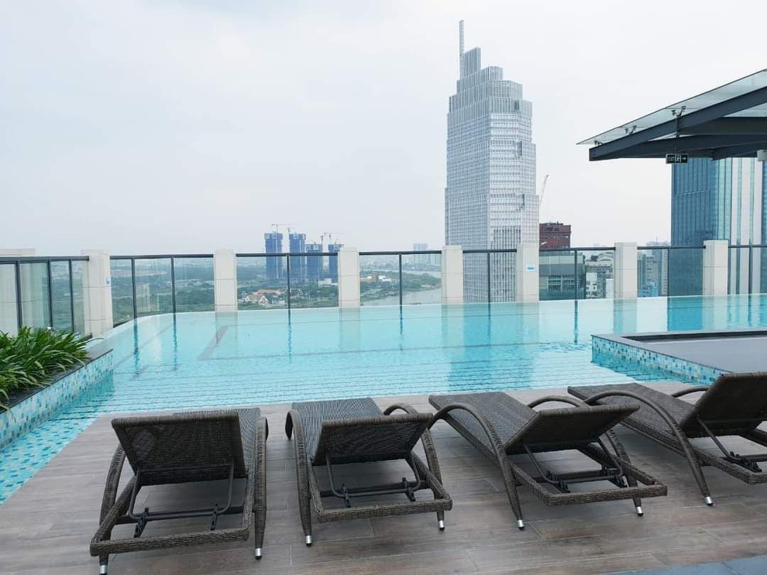 An infinity pool is featured on the rooftop, offering panoramic views of the city skyline. Several lounge chairs are positioned along the edge, creating a relaxed atmosphere. The pool's water surface reflects the clouds above, emphasizing the open and airy environment.