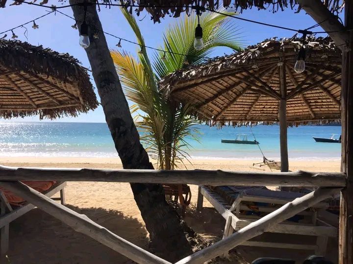 A serene beach scene features traditional thatched umbrellas providing shade on soft sand. Palm trees are visible, swaying gently in the breeze. The tranquil turquoise water extends towards the horizon, where small boats are anchored.