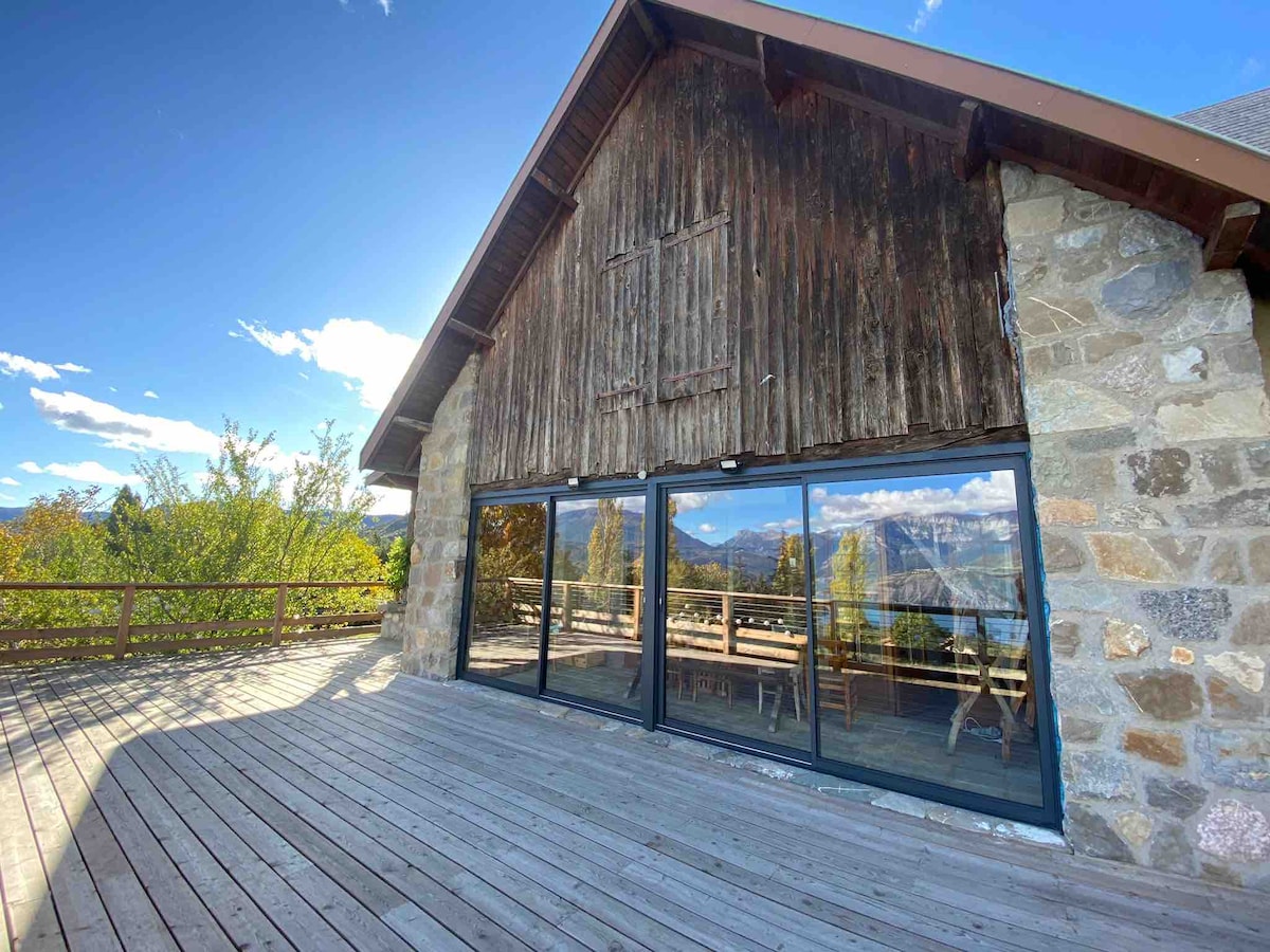 A rustic stone house features large glass doors that open onto a spacious wooden deck. The surrounding landscape includes a clear view of the mountains and the lake, with ample natural light enhancing the outdoor space.