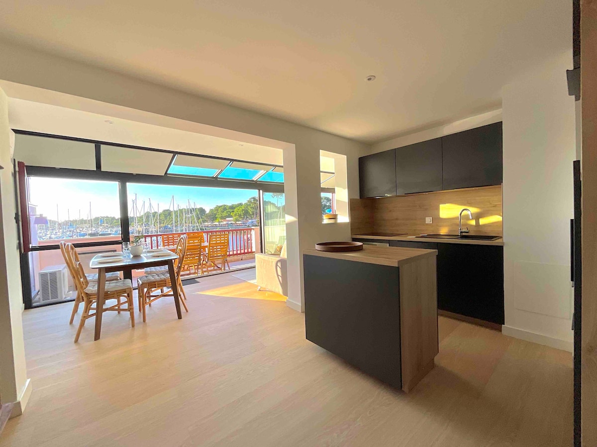 An open-plan living area features a modern kitchen with a sleek black cabinetry and a light wooden island. Natural light fills the space through large windows, offering a view of the harbor. A dining table and chairs are positioned nearby, with access to an outdoor terrace.