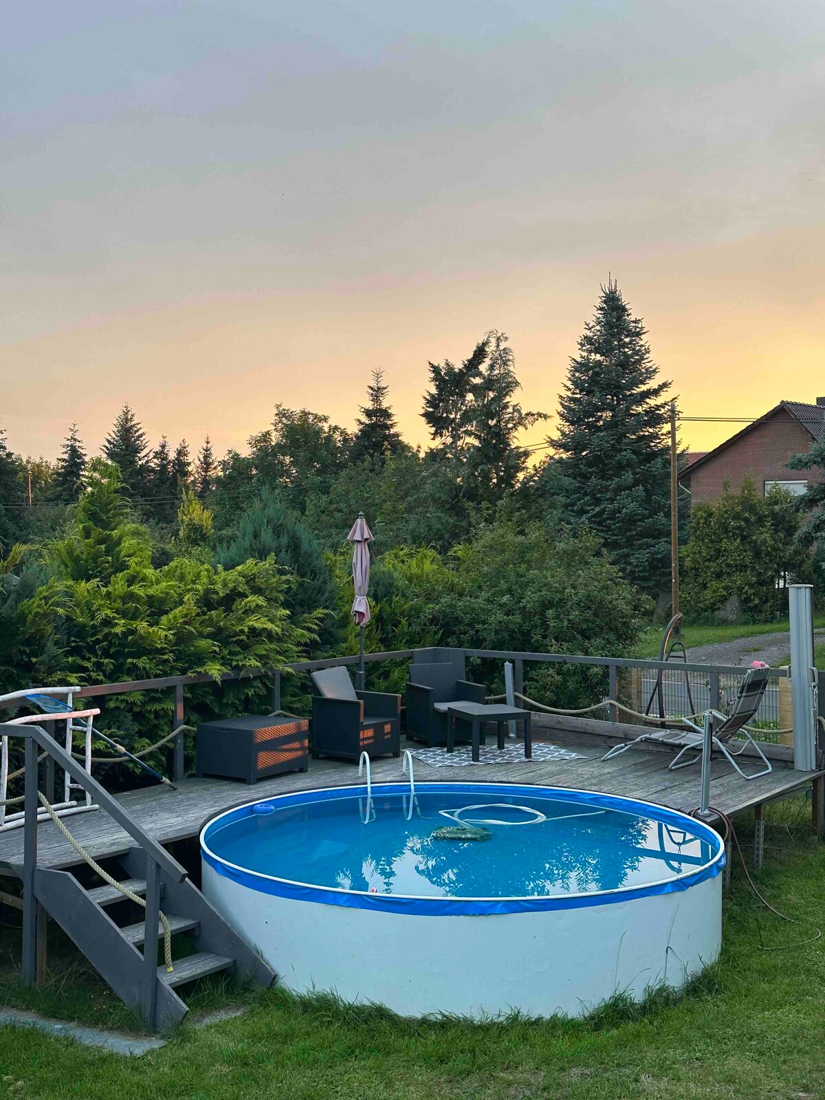 A circular above-ground pool is situated on a wooden deck surrounded by lush greenery. Two black lounge chairs and a small table are positioned nearby, offering a relaxed seating area. The soft colors of the sunset create a serene backdrop to the scene.