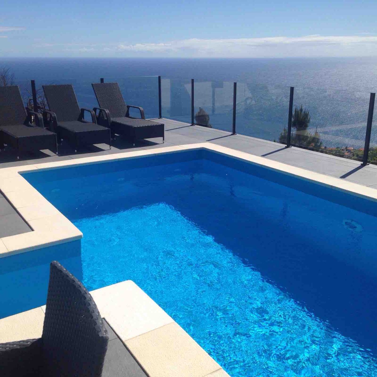A vibrant blue pool is framed by a light-colored stone deck and surrounded by transparent glass railings. Several black lounge chairs are positioned on one side, offering views of the ocean and sky in the background.