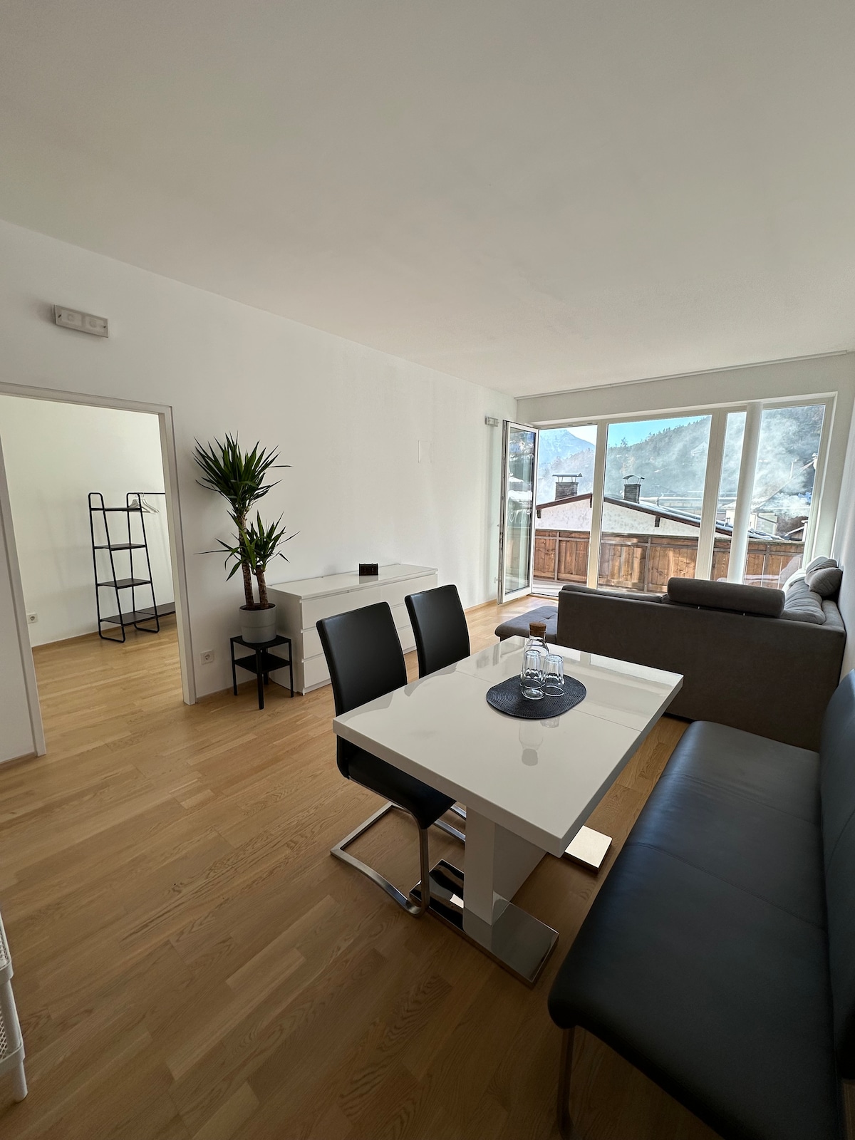 A modern living area is showcased with a sleek white dining table and black chairs. A comfortable gray sofa is positioned nearby, with a large window allowing natural light and offering a view of the outdoor space. A potted plant adds a touch of greenery.