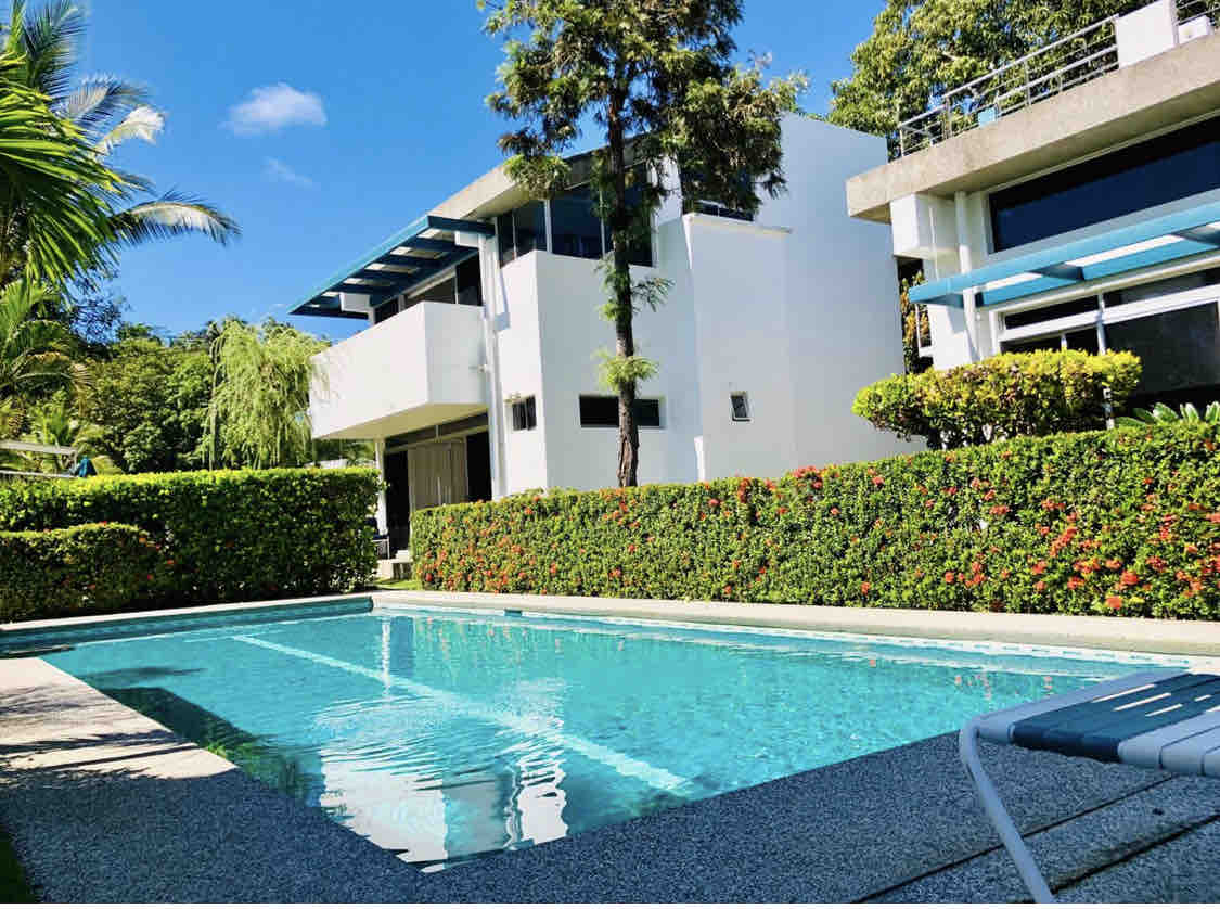 A clear blue swimming pool is surrounded by lush greenery and vibrant flowering bushes. The modern architecture of the adjacent buildings features large windows, accentuating natural light. A poolside lounge chair is positioned nearby, inviting relaxation in the serene outdoor space.