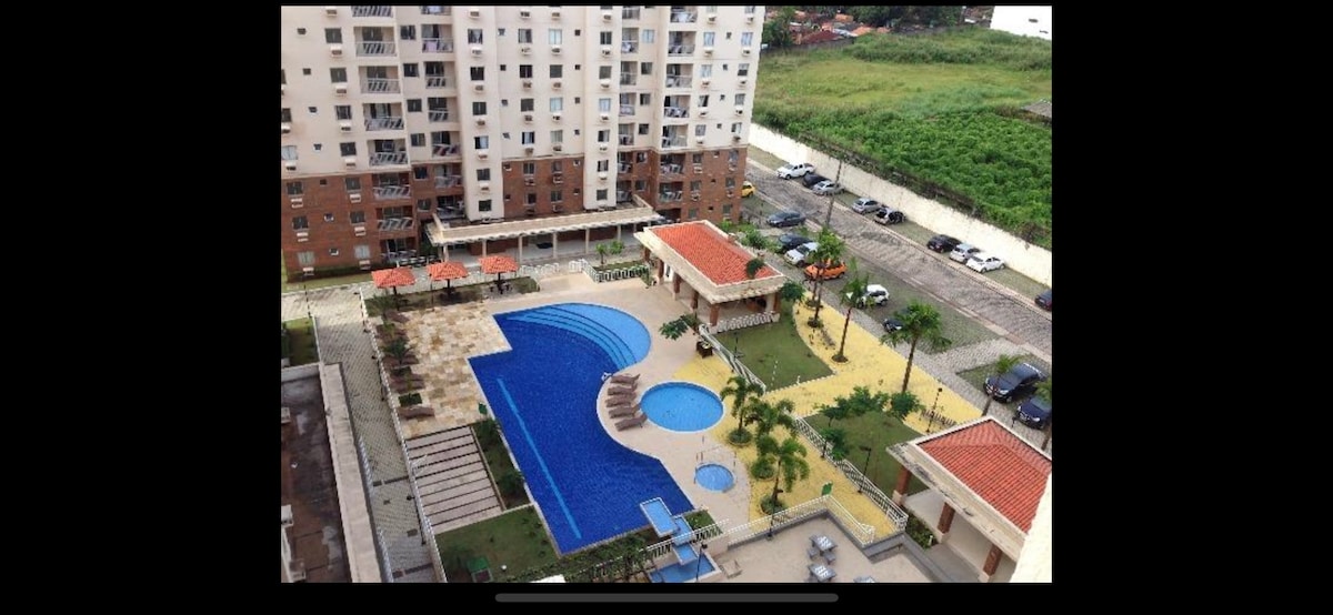 An aerial view captures the expansive condominium grounds featuring two pools, one large and one for children. Surrounding greenery and palm trees enhance the outdoor space, while a covered area with seating provides a gathering spot. Nearby, parking and walkways are visible.