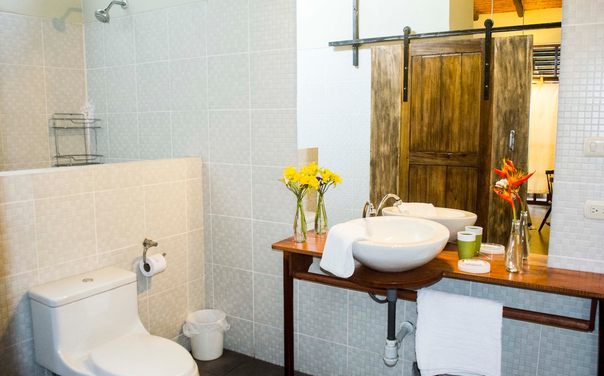 A spacious bathroom features a wooden sliding door, a modern sink set on a wooden countertop, and a wall-mounted mirror. Two flower vases with fresh blooms add a touch of color, while essential toiletries and towels are neatly arranged for convenience.
