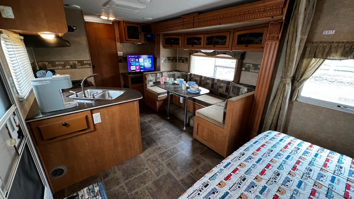 An inviting living space features a well-equipped kitchen area with a sink and stove. A TV is mounted on the wall, and a dining nook with a U-shaped seating arrangement is visible. The cozy bedroom area displays a colorful patterned blanket on the bed.