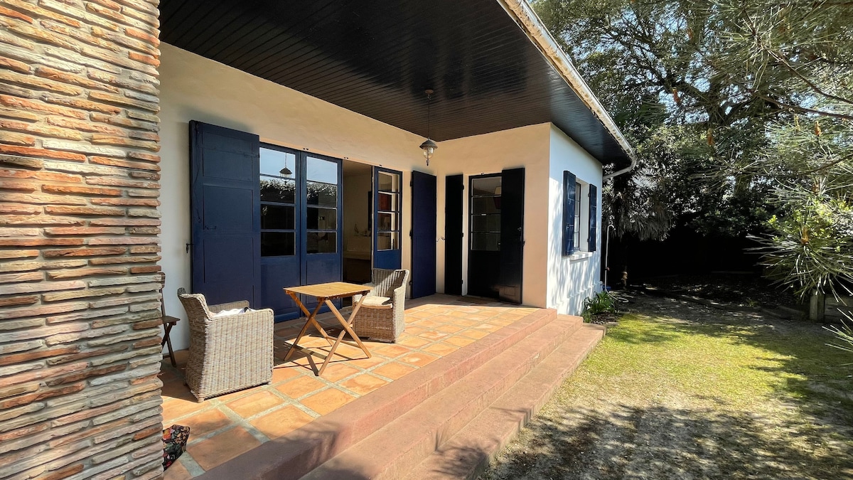 A welcoming outdoor terrace features a rustic wooden table and two wicker chairs, placed on a tiled surface. Blue shutters frame the windows, while a lush garden area with greenery extends beyond. Natural light enhances the inviting atmosphere of the space.