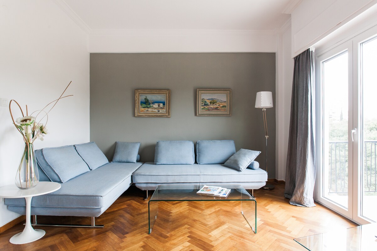 A modern living area features a light blue sectional sofa complemented by a glass coffee table. Artwork is displayed on the wall, and natural light enters through large glass doors that open to a balcony. A floor lamp stands in the corner, enhancing the space's ambiance.