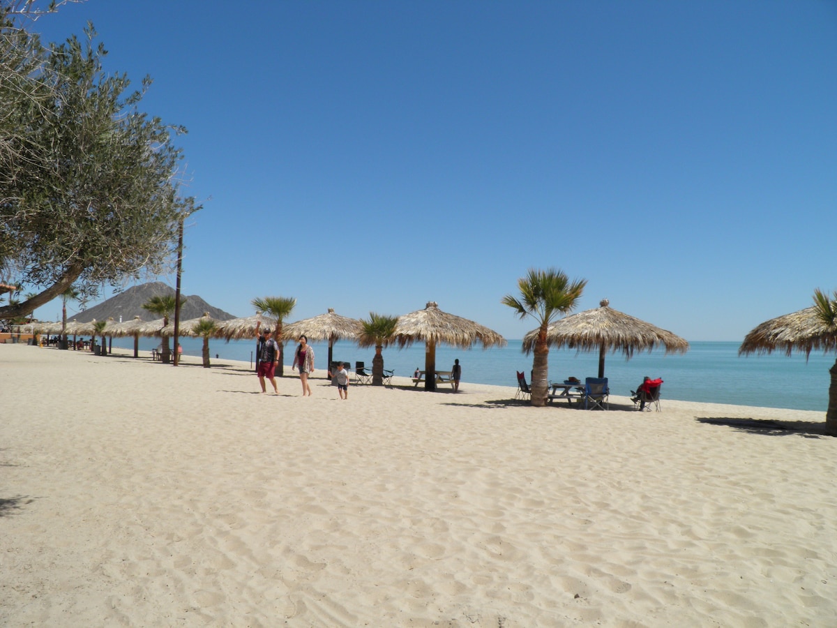A sandy beach stretches under a clear blue sky, with several palapas providing shaded seating areas. People are seen walking along the shore, while others are seated nearby, enjoying the serene view of the calm sea and distant hills.