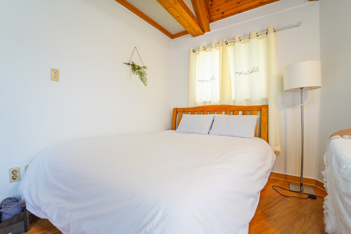 A spacious sleeping area features a wooden bed with a simple white duvet and light blue pillows. Natural light filters through sheer yellow curtains, complementing the warm wooden accents on the ceiling. A standing lamp provides additional illumination, creating a calm environment.