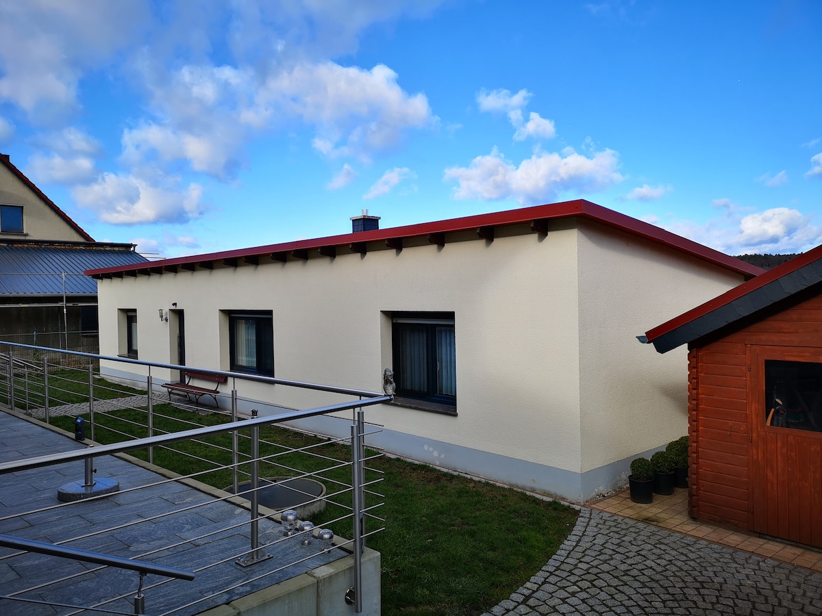 A modern two-room holiday house is presented with a simple exterior, featuring a red roof and white walls. Windows are evenly spaced, allowing natural light. A well-maintained outdoor area includes a terrace pathway and a wooden garden shed nearby, set against a backdrop of a clear blue sky.