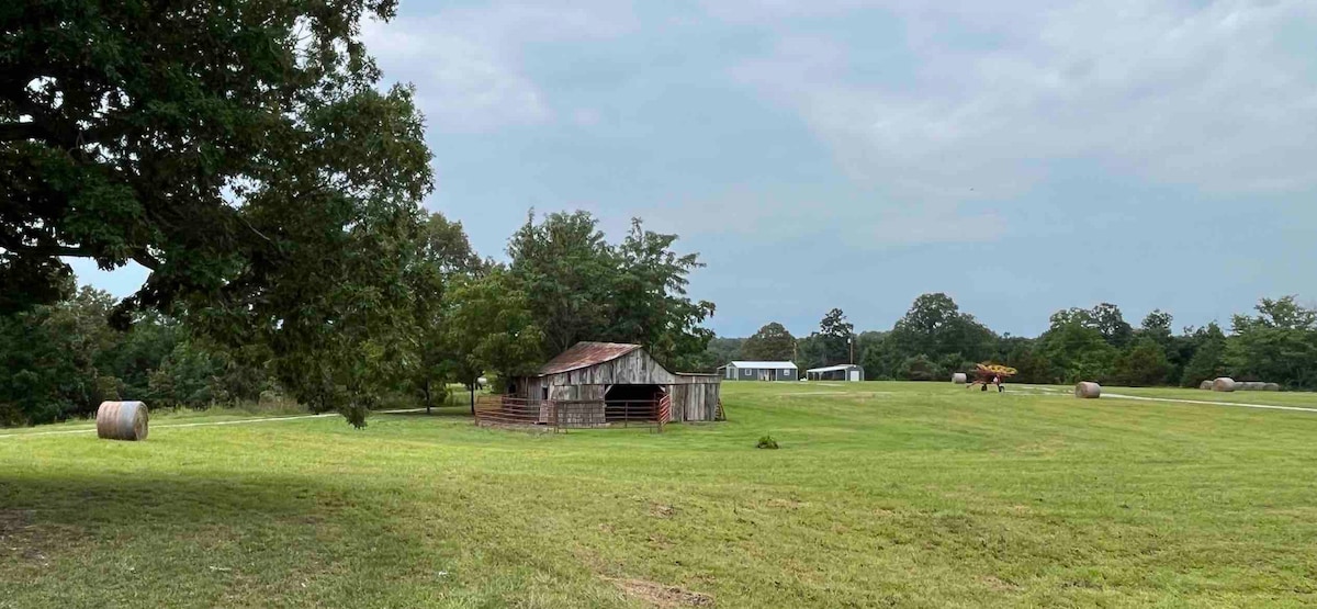 A rustic barn sits in an open field surrounded by green grass and trees. The landscape features distant structures and a few hay bales, with a clear sky overhead, creating a serene rural atmosphere.