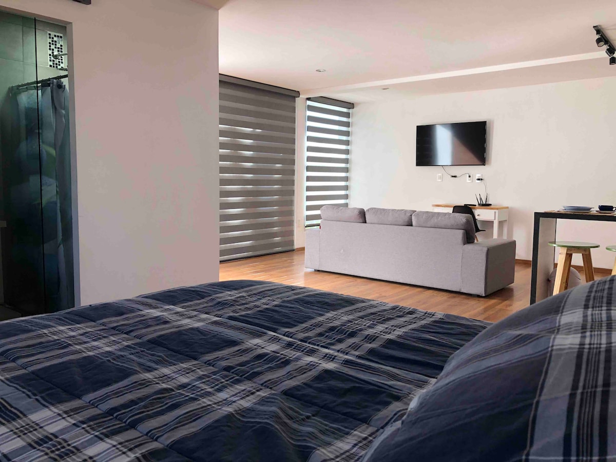 The loft's interior features a cozy sleeping area with a plaid bedspread. A comfortable gray sofa is positioned near a flat-screen TV mounted on the wall. Natural light filters through modern window shades, creating a relaxed atmosphere.