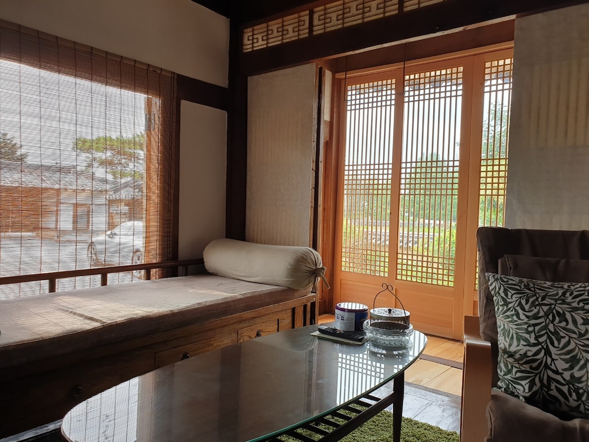 The interior space features a wooden daybed and a glass coffee table, complemented by a green area rug. Sliding doors with intricate lattice designs open to a serene outdoor view, allowing natural light to fill the room. Decorative items are arranged on the table for a welcoming touch.