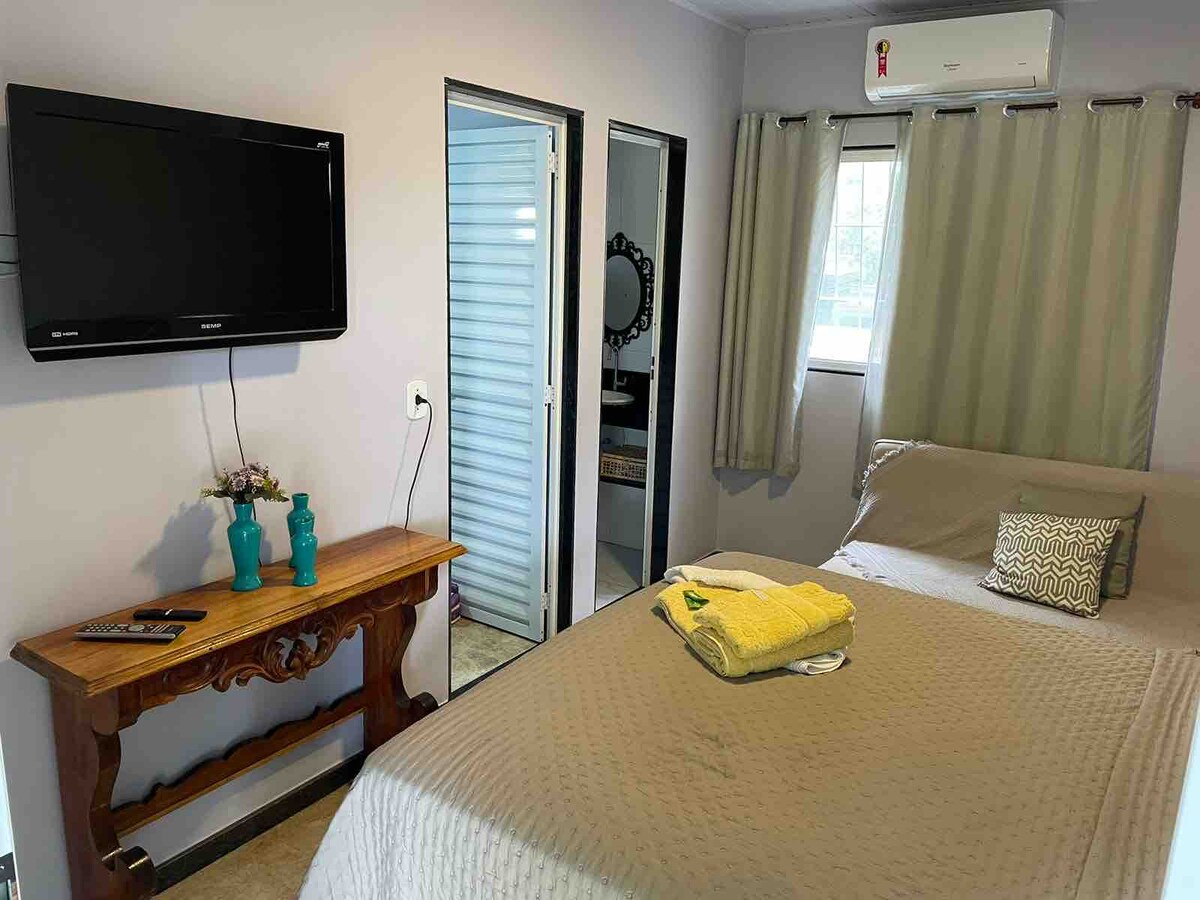 A cozy bedroom is featured, furnished with a spacious bed and a flat-screen television mounted on the wall. Light curtains frame the window, complementing the neutral tones. A decorative table holds two turquoise vases and a folded towel, adding a touch of charm to the space.