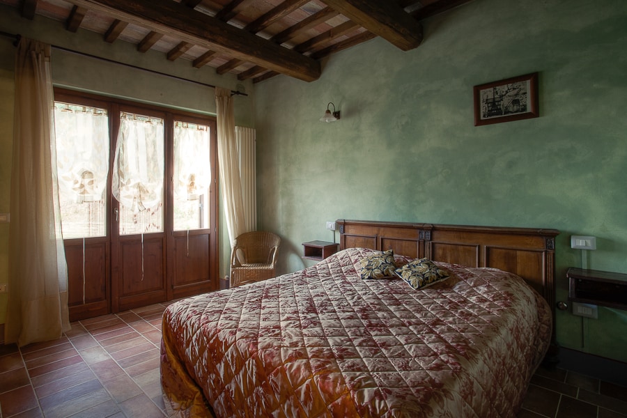 A spacious bedroom features a large bed with a patterned coverlet. Natural light streams through two windows, framed by sheer curtains, illuminating the rustic wooden beams on the ceiling. A wicker chair is positioned near the window, and a wall-mounted light fixture provides additional illumination.
