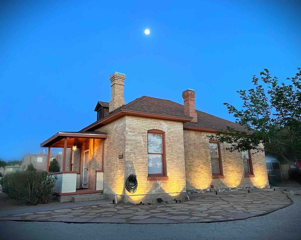 A charming 1940s limestone brick home stands under a twilight sky, enhanced by soft outdoor lighting. The entrance features a welcoming porch with a roof, and a well-maintained pathway made of large stones leads up to the front door. Lush greenery surrounds the property.