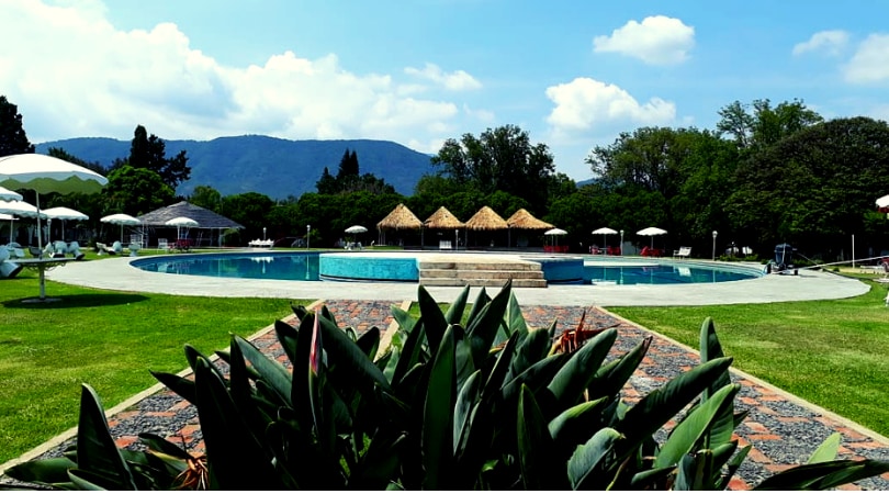 A large recreational pool is set within a sprawling green space, surrounded by lounge chairs and umbrellas. Thatched-roof structures provide shaded seating areas. Lush vegetation enhances the outdoor environment, with distant mountains visible against a partly cloudy sky.