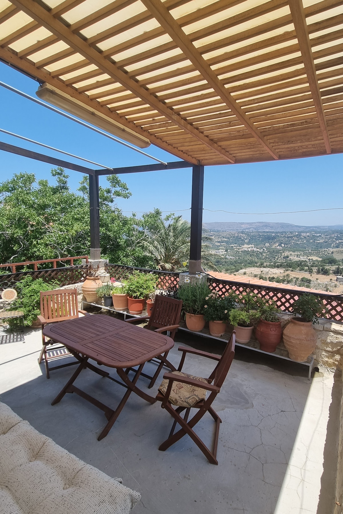 A shaded terrace features a wooden dining table accompanied by several chairs, surrounded by a variety of potted plants. Scenic mountain views can be seen in the background, enhancing the outdoor space's tranquility.