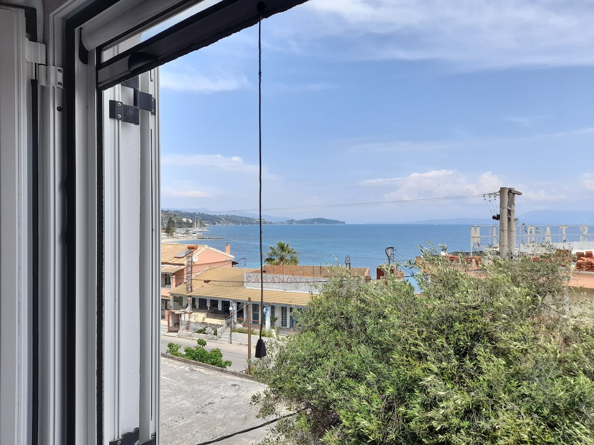 A view from an open window reveals a tranquil coastal scene, showcasing the blue sea under a partly cloudy sky. Buildings line the waterfront, with palm trees adding a touch of greenery, while the horizon softly blends into the sky.