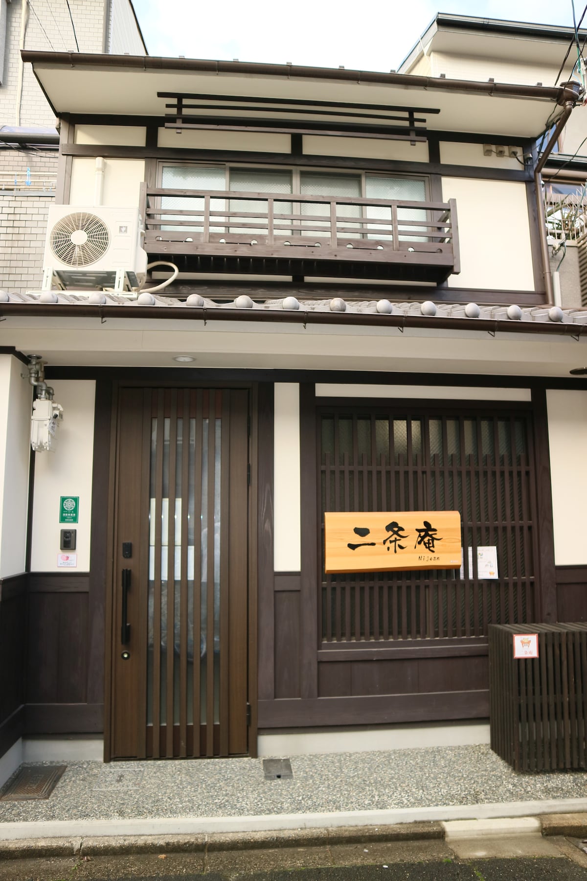 The two-story house features a traditional wooden facade with decorative sliding doors. A wooden sign displaying the name is prominently installed above the entrance. An air conditioning unit is visible on the upper floor, while pebbled pathways lead to the door.