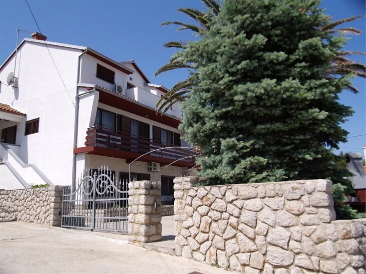 A two-story building is presented, characterized by a white exterior and a red-accented balcony. A well-maintained stone wall and gated entrance frame the scene, complemented by lush green trees that provide shade and environment.