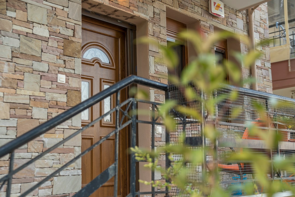 The entrance features a wooden door framed by a textured stone facade. A metal railing leads up a short flight of stairs, while greenery adds a natural touch to the approach, creating a welcoming entrance to the home.