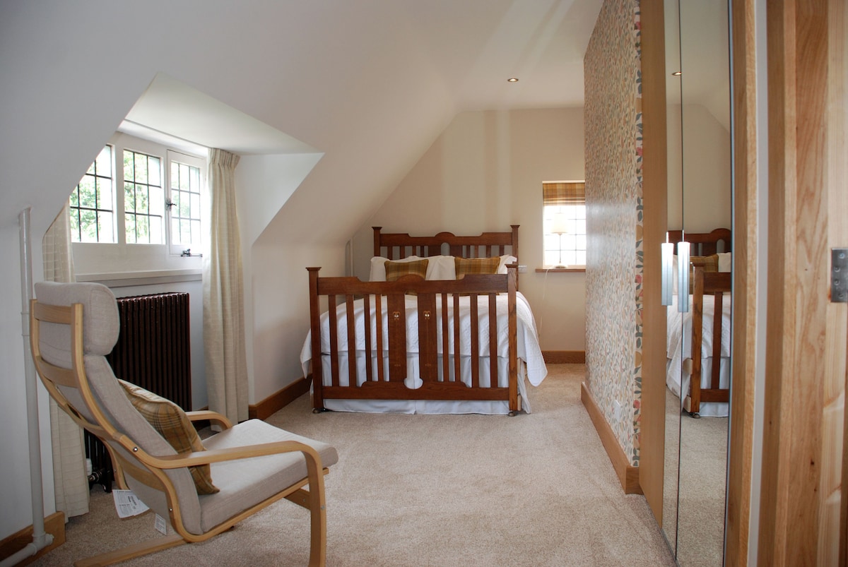 A cozy bedroom features an antique wooden double bed positioned against a light wall. Natural light enters through a leaded window, highlighting soft carpet and serene decor. A comfortable armchair is placed beside the bed, and mirrored wardrobes reflect the room's inviting atmosphere.