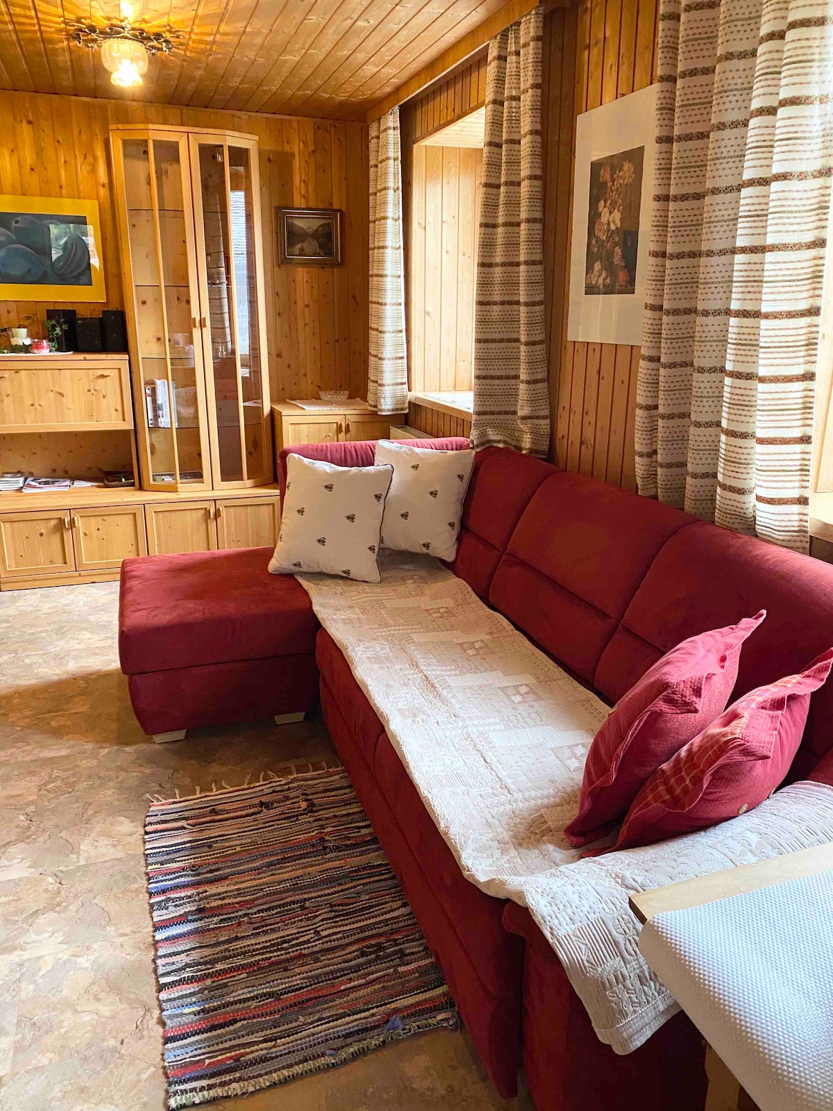 A comfortable red sectional sofa is adorned with several decorative pillows, positioned against a wooden wall. A woven rug lies underneath, while sunlight filters through the windows, illuminating the cozy interior that includes a small television and wooden storage units.