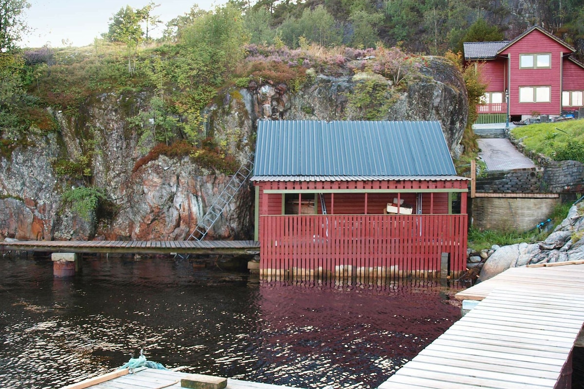 A red boat house with a metal roof is situated at the water's edge, surrounded by rocky terrain and greenery. A wooden dock extends towards the house, and a ladder leads from the dock to the rocky bank. In the background, a larger red house is visible.