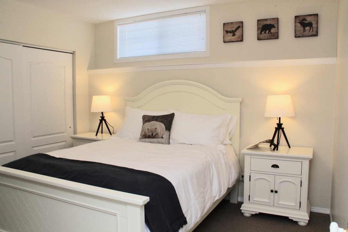 A bright bedroom features a queen size bed with white linens and a dark throw blanket. Two lamps illuminate the space on nightstands beside the bed. Three framed animal images are displayed above the bed, and a window allows natural light to enter.