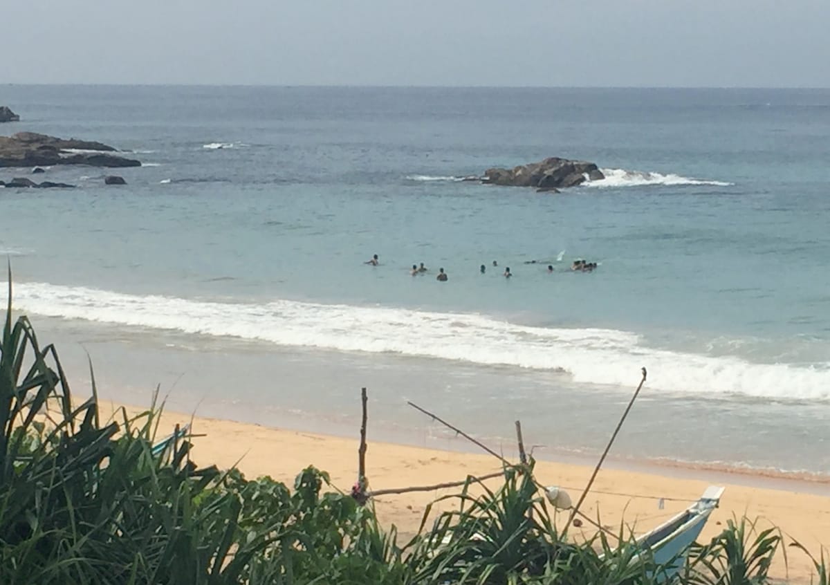 The serene beach scene displays gentle waves lapping the shore, with several people swimming in the clear turquoise water. A sandy beach stretches toward rocky formations, while lush greenery frames the foreground, adding a natural boundary to the coastline.
