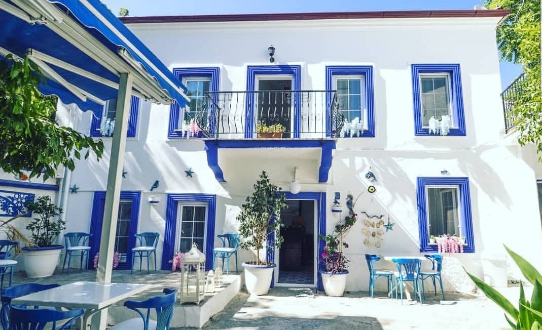 A charming exterior showcases a white building with blue accents, featuring multiple windows and a balcony adorned with potted plants. Outdoor seating is available at white tables and blue chairs, complemented by greenery in pots, creating a welcoming communal space.
