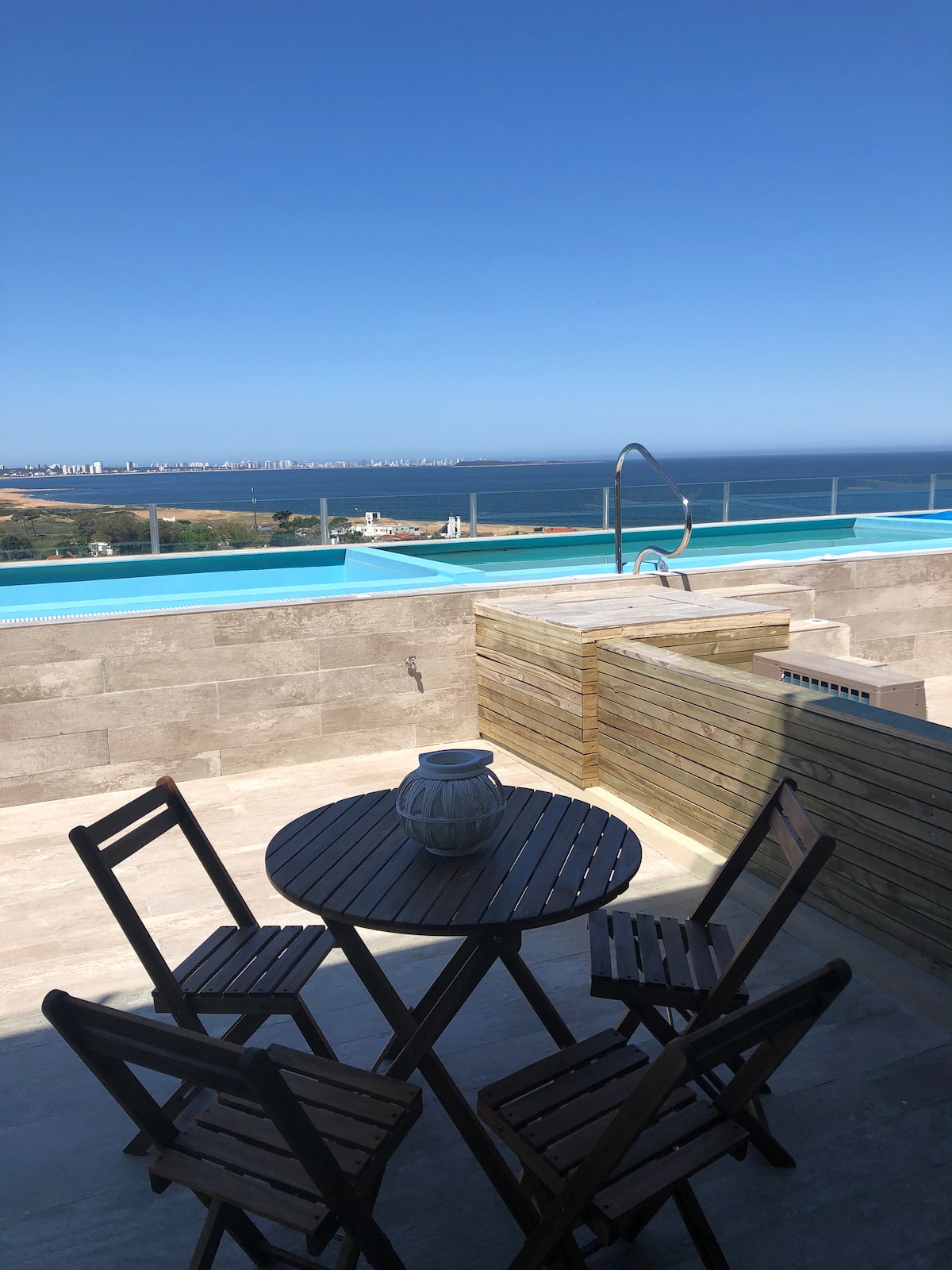 A private terrace is presented, featuring a small round table with four wooden chairs. In the background, a pool offers views of the sea and surrounding landscape, with a clear blue sky above.