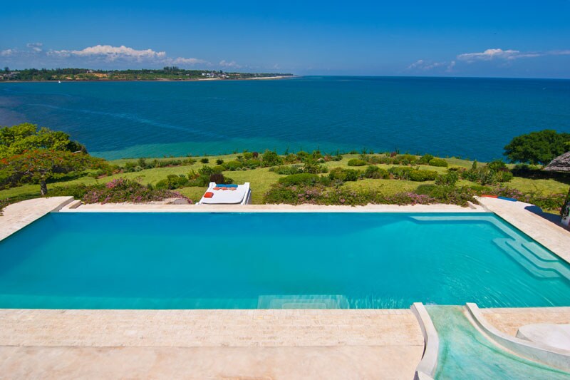 A serene swimming pool is situated at the edge of a beautifully landscaped garden, overlooking the vibrant blue ocean. The tranquil water reflects the clear sky, with sun loungers positioned nearby for relaxation while enjoying sweeping views of the coastline.