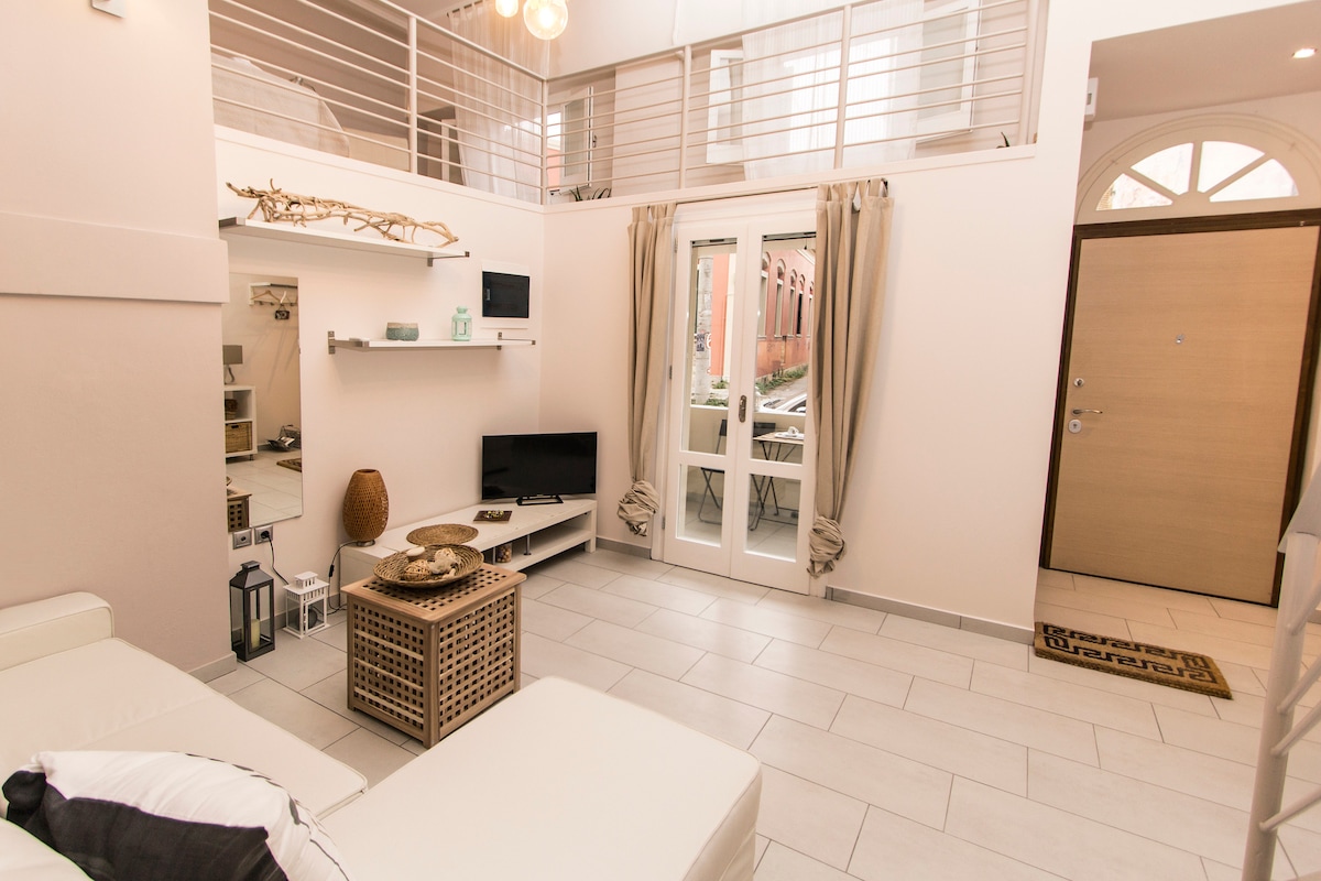 The open-plan living room features a cozy sofa facing a wall-mounted TV. A decorative wooden table is placed in front, and natural light fills the space through large windows with light curtains. The entrance door is visible, leading to the welcoming foyer.