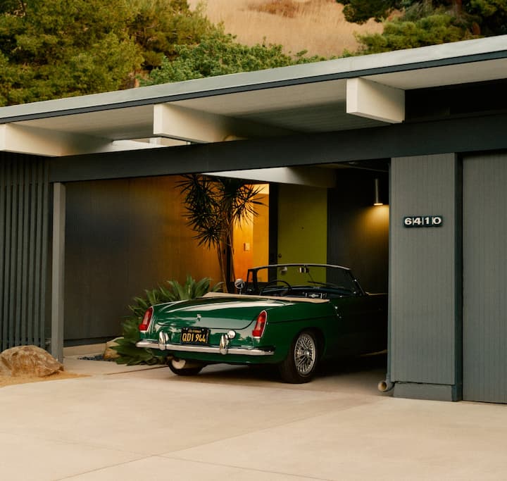 Une photo montre l'extérieur d'une maison de plain-pied dans le désert, dotée d'une place de parking couverte où se trouve une voiture de sport classique décapotable.