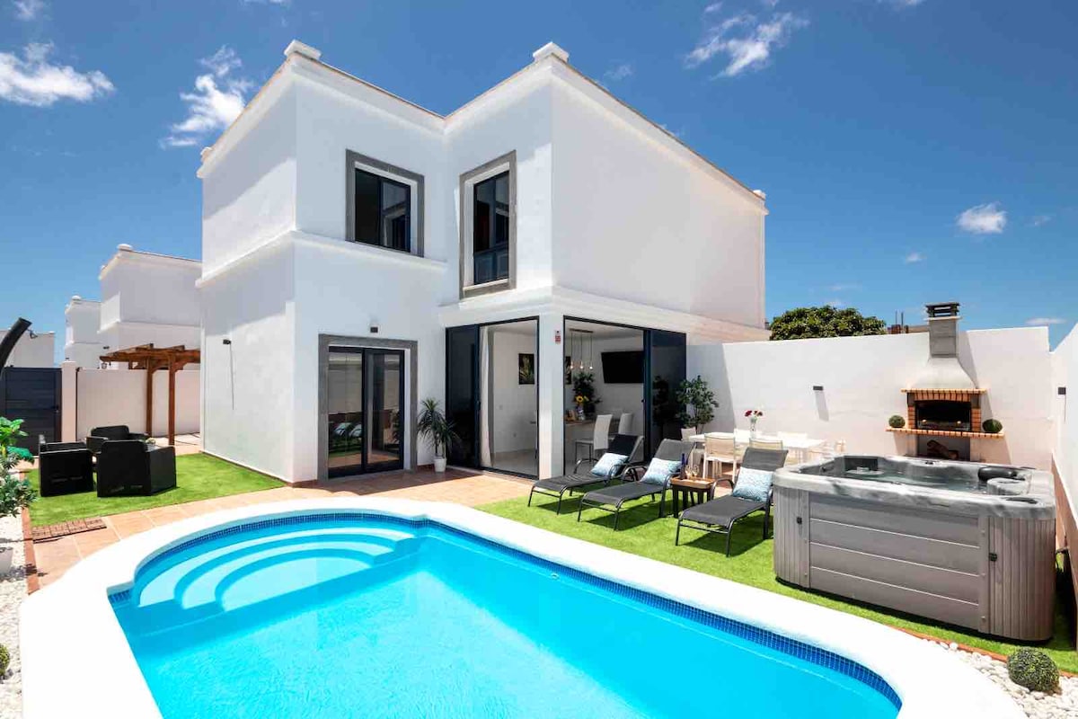 The exterior of the villa showcases a private swimming pool, jacuzzi, and a well-maintained garden area. Comfortable lounge chairs are arranged beside the pool. The house features large glass doors, offering light-filled access to outdoor living spaces, complemented by a patio area with seating.