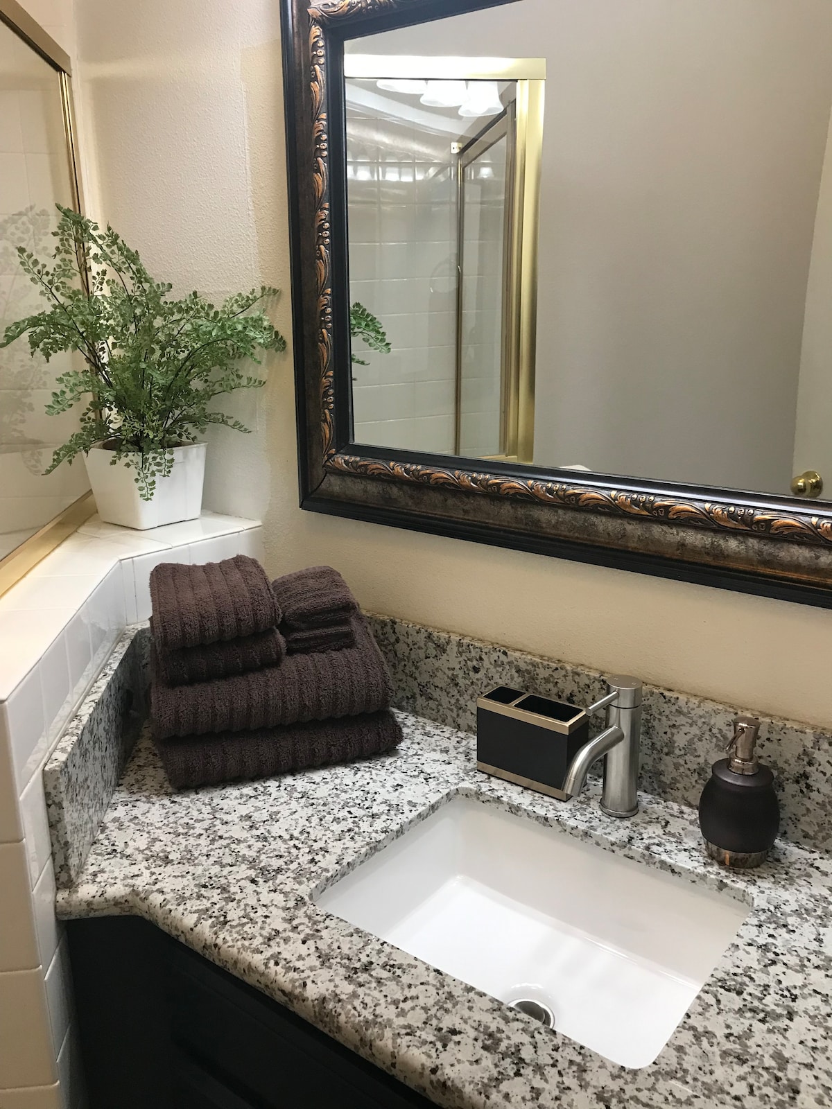A well-appointed bathroom features a marble countertop with neatly arranged dark towels. A decorative plant sits in a white pot above the counter. A large mirror framed in gold reflects the modern fixtures and a shower area is visible in the background.