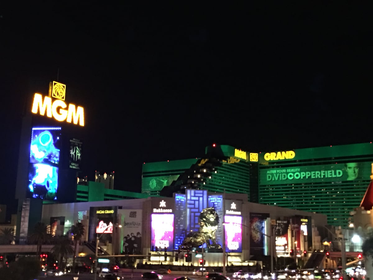 The iconic MGM Grand sign is illuminated against the night sky, along with vibrant digital displays featuring entertainment advertisements. The hotel’s green façade is prominent, showcasing its distinctive architectural design. Bright lights from various attractions create a lively atmosphere reflecting the energy of the Las Vegas Strip.