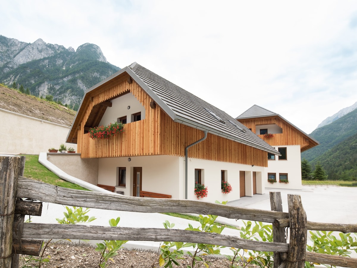 A modern building featuring a charming wooden facade, complemented by flower boxes under the windows. The structure is nestled among rolling hills and mountains, showcasing its natural surroundings. A spacious outdoor area is visible, accommodating parking and easy access.