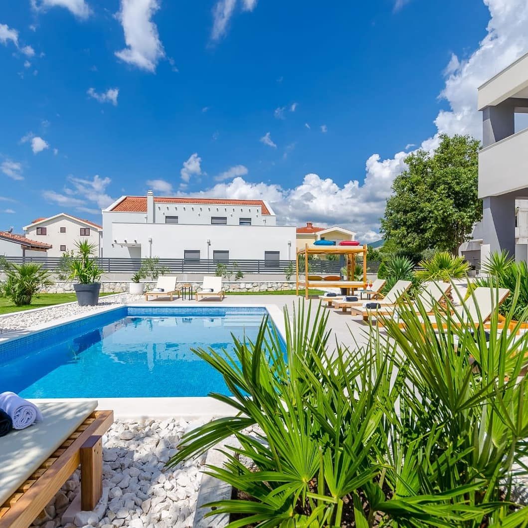 A serene outdoor space features a rectangular swimming pool surrounded by tropical plants and sun loungers. A shaded seating area is visible, providing a relaxed atmosphere. The clear blue sky and scattered clouds enhance the inviting setting, ideal for leisurely afternoons.