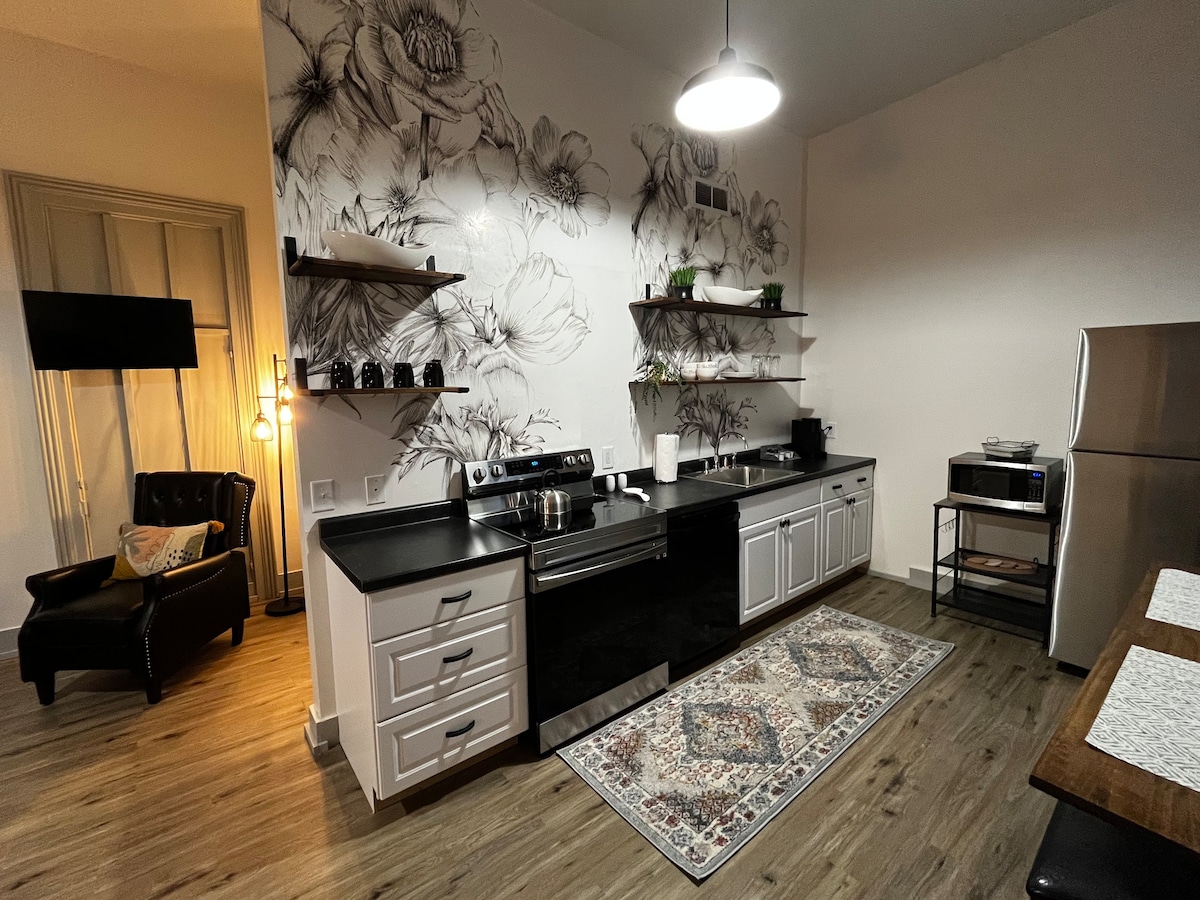 A modern kitchen area features sleek black appliances and white cabinetry, complemented by a decorative floral wall mural. Open shelving displays various kitchen items, while a small table sits nearby. A cozy armchair in the corner provides seating, illuminated by a pendant light.