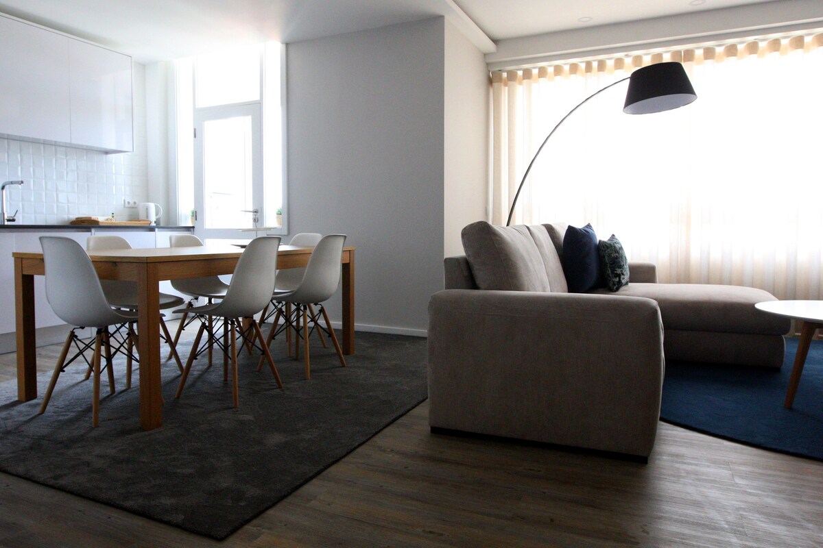 An open living area features a dining table with white chairs and a comfortable beige sectional sofa. A striking floor lamp adds visual interest. Natural light filters through sheer curtains, creating a warm and bright atmosphere in the space.