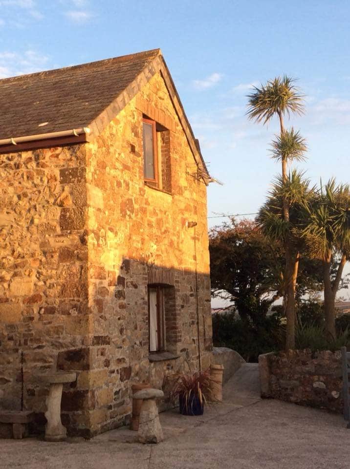 The exterior of a charming stone barn-style building is highlighted by warm sunlight. Lush green palm trees sway nearby, complementing the natural landscape. A gravel pathway leads to the entrance, framed by stone features and terracotta pots, creating an inviting outdoor area.