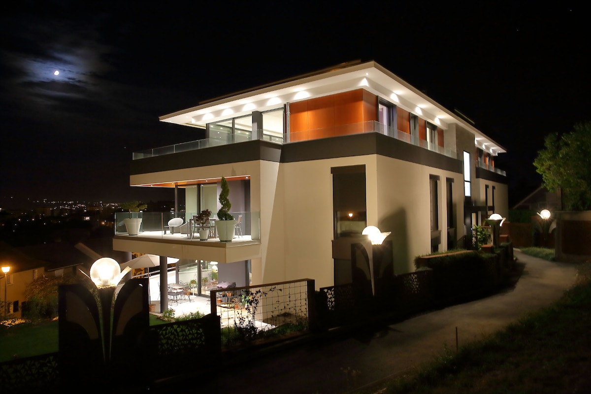 The modern building is showcased at night, illuminated by soft outdoor lighting. A spacious balcony extends from the upper floor, offering views of the surrounding area. The landscape is gently sloped, lined with decorative lamps and greenery, enhancing the tranquil evening scene.